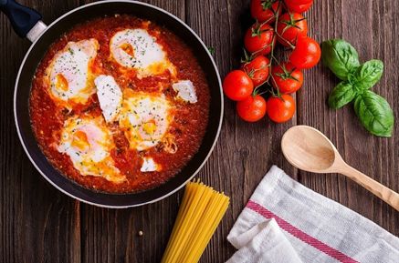 Shakshuka dish with poached eggs and tomatoes in a skillet, surrounded by a branch of tomatoes, a wooden spoon and a tea towel on a wooden table.