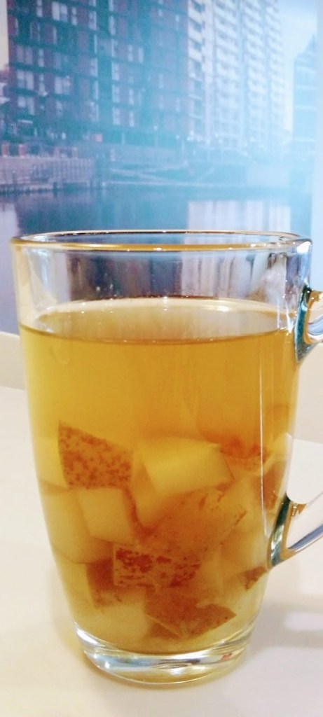 A transparent mug of ginger pear tea (with chunks of pear inside) on a white breakfast table, with a picture of a scene showing buildings around a lake in the background, which can be seen just above the mug.