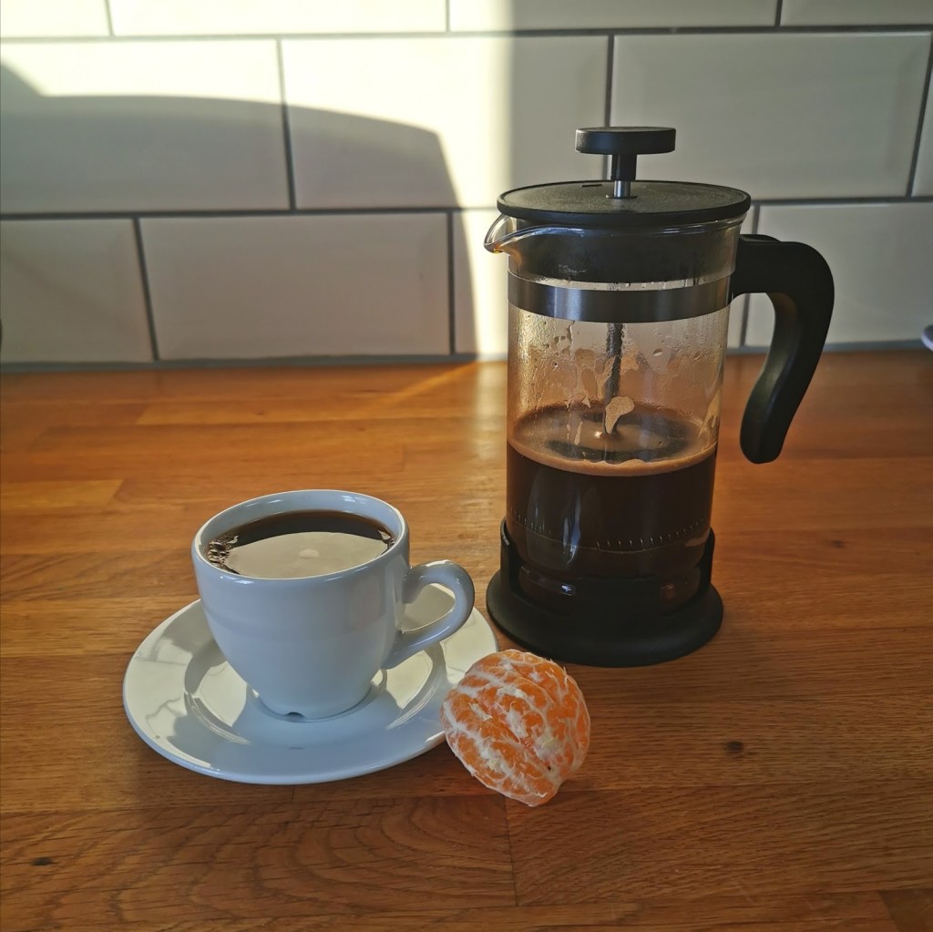 A cup of coffee on a small plate with a clementine segment in front of it, to the right, sitting on a wooden kitchen table. A coffee strainer is next to the cup.