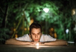 A guy looking at a candle in the dark, against a green backdrop.