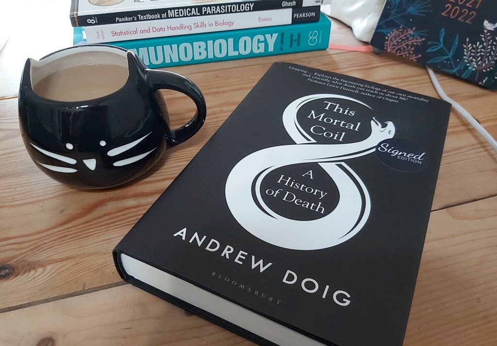 Andrew Doig's 'This Mortal Coil: A History of Death' book on a table, next to a black cat mug and a stack of science books.