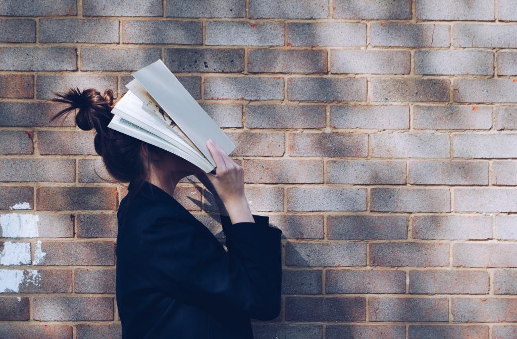 A lady with her hair tied in a bun, wearing a black suit, placing an open white book over her face (facing side on to both a wall and the viewer). 